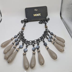 Könnte beinhalten: Eine Mixit-Halskette mit einer dunklen Kette aus Gunmetal und tropfenförmigen Anhängern. Die Halskette hat graue und klare Perlen, mit dem Markennamen "MIXIT."