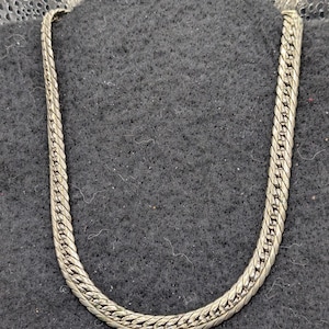 May include: A silver-toned metal chain necklace featuring a flat, interlocking herringbone-style link design. The chain has a smooth, reflective surface and is arranged in a U-shape, showcasing the consistent pattern of the metallic links throughout.