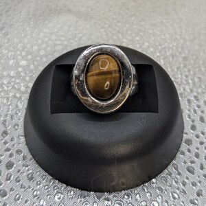 May include: A silver-toned ring featuring an oval tiger's eye stone. The stone is a warm brown color with natural patterns, set within a textured silver bezel. The ring is displayed on a black pedestal.