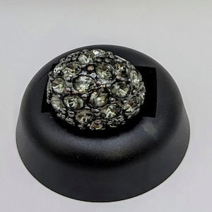 Può includere: Un anello argentato con un gruppo di pietre chiare e sfaccettate. L'anello è esposto su un supporto nero su sfondo bianco. Il design dell'anello presenta una forma a cupola, con le pietre molto vicine.