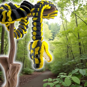 May include: A segmented, articulated snake toy featuring a vibrant pattern of alternating black and bright yellow sections. The snake is coiled around a brown, textured branch-like stand, with its head hanging downward and mouth slightly open.