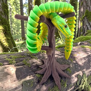 May include: A 3D-printed articulated snake in shades of green and yellow, coiled around a miniature brown tree. The snake is resting on a moss-covered log, with a forest background.