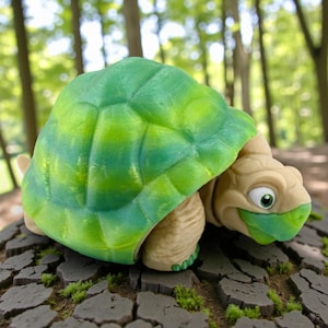 May include: A whimsical, cartoon-style turtle figurine. The turtle has a green and yellow shell, tan body, and a friendly expression. It is posed on a textured, dark gray surface with patches of green moss, set against a blurred forest background.