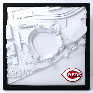 May include: A framed 3D white relief map of a baseball stadium and surrounding city infrastructure, featuring a prominent red, white, and black Reds logo in the bottom right corner, all encased within a simple black square frame.