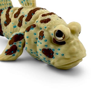 May include: A 3D printed articulated mudskipper fish figurine featuring a cream-colored body with brown patches and small bright blue spots. The fish has large, prominent eyes and a segmented body structure that allows for flexible movement.
