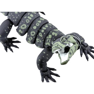 May include: An articulated lizard figurine featuring a dark charcoal gray body with light green circular and irregular spot patterns. The creature has a textured skin surface, four splayed feet with distinct toes, and an open mouth showing white.