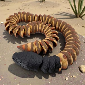 May include: A detailed, segmented sculpture of a snake, featuring a black head and a body with alternating bands of brown and tan. The snake is coiled on a sandy surface, with a few plants in the background.