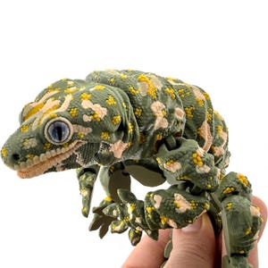 May include: An articulated gecko figurine featuring a textured olive green body with mottled patches of pale cream and bright yellow. The lizard has large, prominent eyes with dark pupils and a detailed, segmented body structure with visible joints.