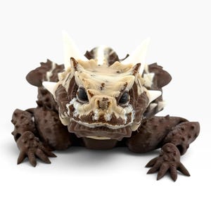May include: A 3D printed horned lizard figurine featuring a dark brown body with a textured, bumpy surface. The head is cream-colored with prominent, sharp, white spikes extending from the sides and top, and dark, glossy, round eyes.