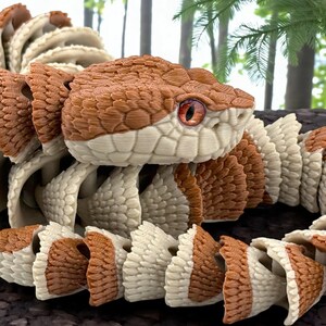 May include: A 3D printed articulated snake figure featuring a repeating pattern of brown and cream-colored segments. The snake has a detailed head with textured scales and a prominent orange eye, showcasing a segmented body design with visible layers.