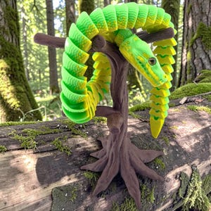 May include: A bright neon green and yellow articulated snake toy coiled around a brown, textured tree-shaped stand. The snake features a segmented body with a detailed head, visible eyes, and a flexible design that wraps around the branches.