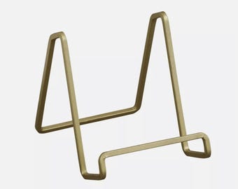 Puede incluir: Un caballete de metal dorado para exhibir fotos, obras de arte u otros objetos pequeños.