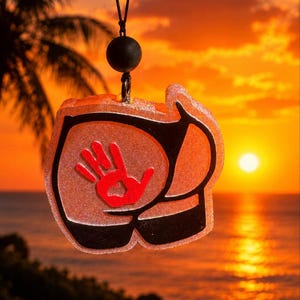 May include: A translucent, glittery orange resin pendant shaped like buttocks wearing black thong underwear, featuring a prominent red handprint graphic. The pendant hangs from a black cord with a single round black bead above it.