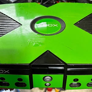 May include: A green and black Xbox console. The console has a large green X on top, with the word "XBOX" in white. The front of the console has buttons and ports. The console is sitting on a black surface.