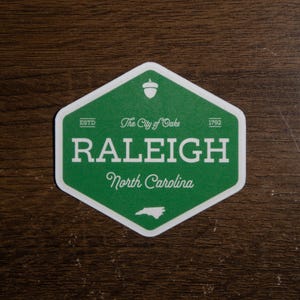 Pu&ograve; includere: Un adesivo esagonale verde con bordo bianco che riporta il testo RALEIGH, North Carolina e The City of Oaks. Include un'icona a forma di ghianda in alto, il testo ESTD 1792 e una piccola sagoma bianca dello stato della Carolina del Nord in basso.