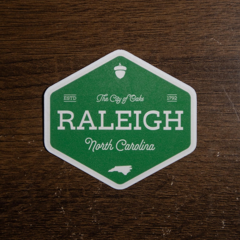 Pu&ograve; includere: Un adesivo esagonale verde con bordo bianco che riporta il testo RALEIGH, North Carolina e The City of Oaks. Include un'icona a forma di ghianda in alto, il testo ESTD 1792 e una piccola sagoma bianca dello stato della Carolina del Nord in basso.