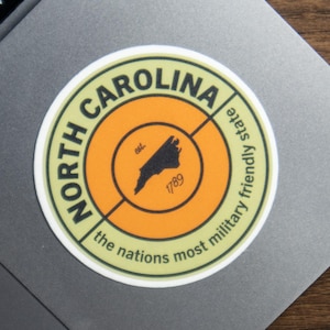 May include: A circular sticker with an orange center and a white border. The text reads NORTH CAROLINA and the nations most military friendly state. In the center is a black silhouette of North Carolina with the text est. 1789.