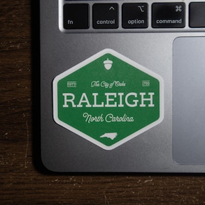 Pu&ograve; includere: Un adesivo esagonale verde con bordo bianco che riporta il testo "The City of Oaks", "RALEIGH", "North Carolina", "ESTD 1792", un'icona a forma di ghianda e una piccola sagoma dello stato della Carolina del Nord, applicato su un portatile.