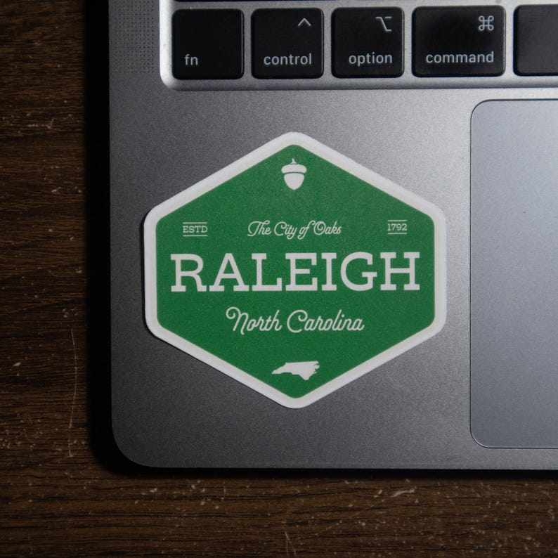 Pu&ograve; includere: Un adesivo esagonale verde con bordo bianco che riporta il testo "The City of Oaks", "RALEIGH", "North Carolina", "ESTD 1792", un'icona a forma di ghianda e una piccola sagoma dello stato della Carolina del Nord, applicato su un portatile.