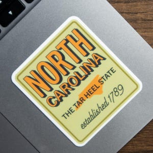 May include: A rectangular sticker with a light yellow background and a white border. The text reads NORTH CAROLINA in bold orange letters, followed by THE TAR HEEL STATE and established 1789 in black font, all centered on the sticker.