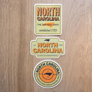 May include: Three rectangular and circular stickers featuring North Carolina themes. The top sticker is rectangular with orange text reading NORTH CAROLINA, THE TAR HEEL STATE, established 1789. The middle is a shaped sign with similar text. The bottom is circular.