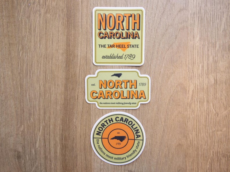 May include: Three rectangular and circular stickers featuring North Carolina themes. The top sticker is rectangular with orange text reading NORTH CAROLINA, THE TAR HEEL STATE, established 1789. The middle is a shaped sign with similar text. The bottom is circular.