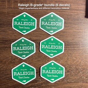 Pu&ograve; includere: Un pacchetto di sei adesivi esagonali verdi con il testo Raleigh North Carolina, The City of Oaks e una piccola icona di ghianda. Il testo Raleigh B-grade bundle (6 decals) *Slight imperfections and different lamination material &egrave; visibile.