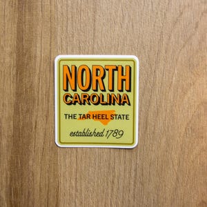 May include: A square sticker with a light olive green background featuring the text NORTH CAROLINA in bold orange letters with black drop shadows. Below is the text THE TAR HEEL STATE with an orange map outline and established 1789 in script.