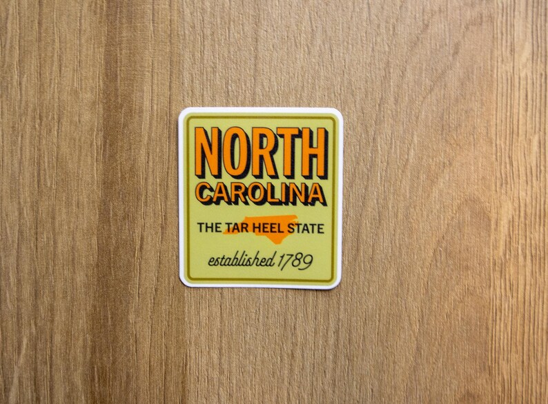 May include: A square sticker with a light olive green background featuring the text NORTH CAROLINA in bold orange letters with black drop shadows. Below is the text THE TAR HEEL STATE with an orange map outline and established 1789 in script.