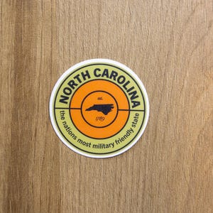 May include: A circular sticker featuring a light green border with the text North Carolina and the nations most military friendly state. The center is orange with a black outline of the state, the text est. and the year 1789 printed in black.
