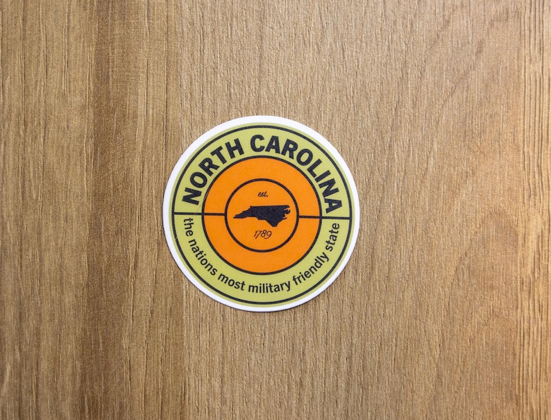 May include: A circular sticker featuring a light green border with the text North Carolina and the nations most military friendly state. The center is orange with a black outline of the state, the text est. and the year 1789 printed in black.