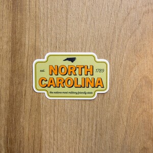 May include: A rectangular sticker with rounded corners featuring a light olive green background. It displays the text 'NORTH CAROLINA' in bold orange letters with black outlines, 'est. 1789', a small black state silhouette, and 'the nations most military friendly state'.