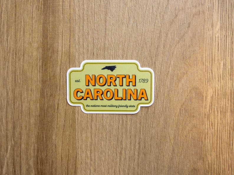 May include: A rectangular sticker with rounded corners featuring a light olive green background. It displays the text 'NORTH CAROLINA' in bold orange letters with black outlines, 'est. 1789', a small black state silhouette, and 'the nations most military friendly state'.