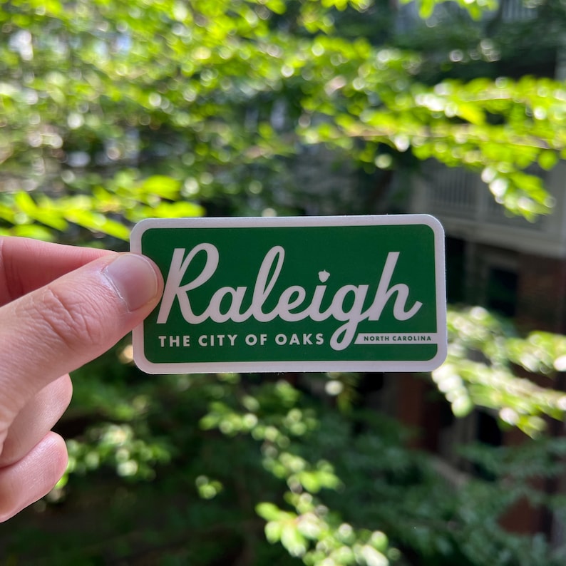 Pu&ograve; includere: Un adesivo rettangolare verde con un bordo bianco che presenta il testo in corsivo bianco Raleigh e il testo in stampatello bianco THE CITY OF OAKS NORTH CAROLINA. L'adesivo ha un design piatto con una tipografia chiara e ben leggibile.