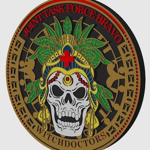 May include: A round, black and gold patch with the words "Joint Task Force Bravo Witchdoctors" around the edge. In the center is a white skull wearing a green, yellow, and red headdress with a red cross on the front.