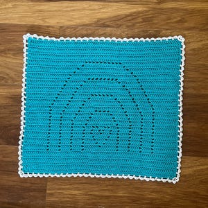 May include: A square, turquoise crochet blanket with a white scalloped border. The blanket features a rainbow design in the center. The handmade blanket is displayed on a wooden surface.