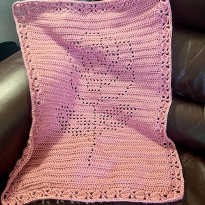 May include: A handmade, pink crochet blanket with a floral design in the center. The blanket features a detailed border and is made of soft yarn. The blanket is approximately 30 inches by 30 inches.