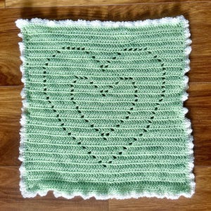 Op de afbeelding: Een vierkant, handgemaakt gehaakt kleed in een lichtgroene kleur met een witte geschulpte rand. Het kleed heeft een hartvormig ontwerp in het midden, gemaakt met open steken. Het kleed is gemaakt van zacht garen.