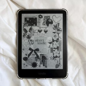 Könnte beinhalten: Ein schwarzer Kindle-E-Reader mit einer transparenten Schutzhülle. Der Bildschirm zeigt eine Collage von Aufklebern, darunter Sterne, Herzen und eine Schleife. Der Text "alexis's kindle" ist sichtbar. Der E-Reader liegt auf weißem, zerknittertem Stoff.