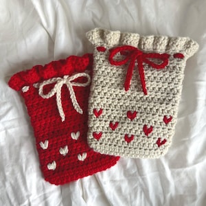Puede incluir: Dos bolsas hechas a mano con ganchillo. Una es roja con corazones blancos y un lazo blanco. La otra es blanquecina con corazones rojos y un lazo rojo. Ambas tienen un borde superior con volantes.