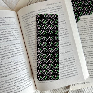 Puede incluir: Marcapáginas negro con un patrón repetido de varitas rosas, sombreros de bruja verdes y coronas doradas. El marcapáginas es rectangular y está sobre un libro abierto.