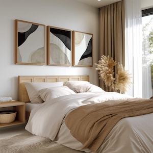 Könnte beinhalten: Ein Schlafzimmer mit einem Bett mit einem hellbraunen Kopfteil und einer weißen und beigefarbenen Bettdecke. An der Wand über dem Kopfteil hängen drei abstrakte Schwarzweißbilder in Rahmen. Eine Vase mit getrocknetem Pampasgras steht auf dem Nachttisch neben dem Bett.