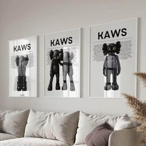 May include: Three black and white framed prints with the word "KAWS" in bold white letters. Each print features a different black and white illustration of a cartoon character with the text "KAWS" in bold white letters.