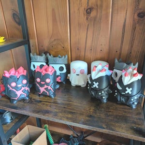 May include: A collection of small, stylized figurines arranged on a shelf. The group includes two black figures with pink crystal-like tops, two dark grey skull-themed figures, one white ghost-like figure with red spiral eyes, and two black figures with white X-shaped eyes.