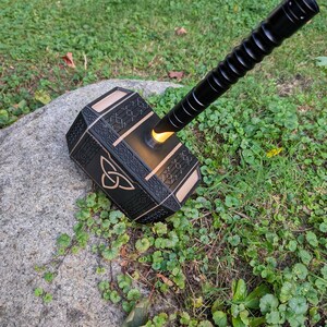 Puede incluir: Un martillo negro y dorado con un mango texturizado y un centro amarillo brillante. El martillo est&aacute; apoyado en una roca en una zona de c&eacute;sped. La cabeza del martillo presenta un dise&ntilde;o de nudo celta.