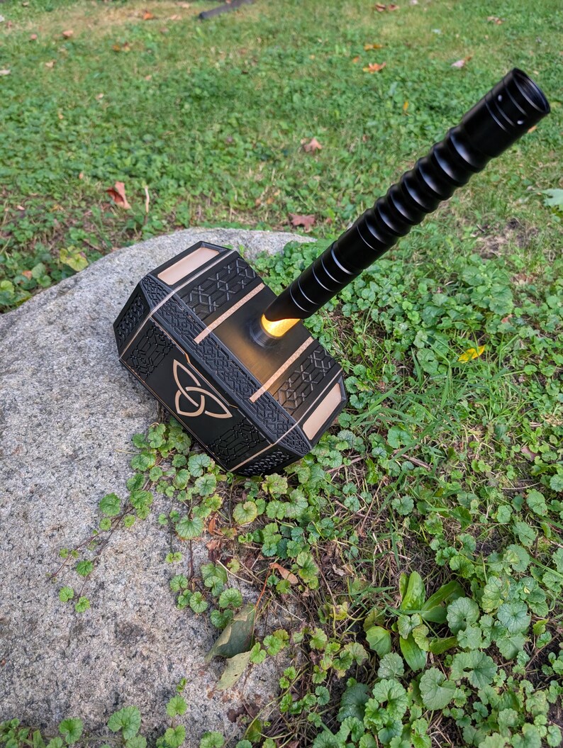 Puede incluir: Un martillo negro y dorado con un mango texturizado y un centro amarillo brillante. El martillo est&aacute; apoyado en una roca en una zona de c&eacute;sped. La cabeza del martillo presenta un dise&ntilde;o de nudo celta.