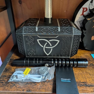 Puede incluir: Un martillo decorativo negro y plateado con un cilindro transparente en la parte superior, un sable de luz negro y un manual del producto. El martillo presenta un dise&ntilde;o de nudo celta. Los accesorios incluyen un cable de carga y un objeto redondo peque&ntilde;o.