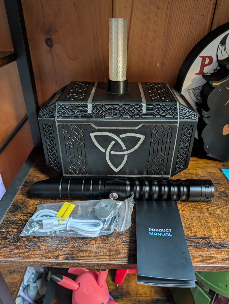 Puede incluir: Un martillo decorativo negro y plateado con un cilindro transparente en la parte superior, un sable de luz negro y un manual del producto. El martillo presenta un dise&ntilde;o de nudo celta. Los accesorios incluyen un cable de carga y un objeto redondo peque&ntilde;o.