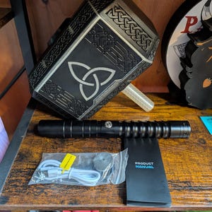 Puede incluir: Una r&eacute;plica negra y plateada del martillo de Thor, Mjolnir, con un dise&ntilde;o de nudo celta. Tambi&eacute;n se ven un sable de luz negro y un manual del producto. Incluye un cable de carga y un peque&ntilde;o disco.