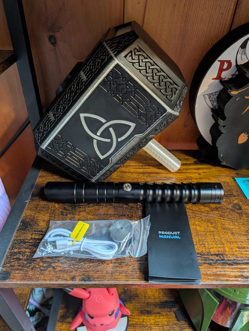 Puede incluir: Una r&eacute;plica negra y plateada del martillo de Thor, Mjolnir, con un dise&ntilde;o de nudo celta. Tambi&eacute;n se ven un sable de luz negro y un manual del producto. Incluye un cable de carga y un peque&ntilde;o disco.